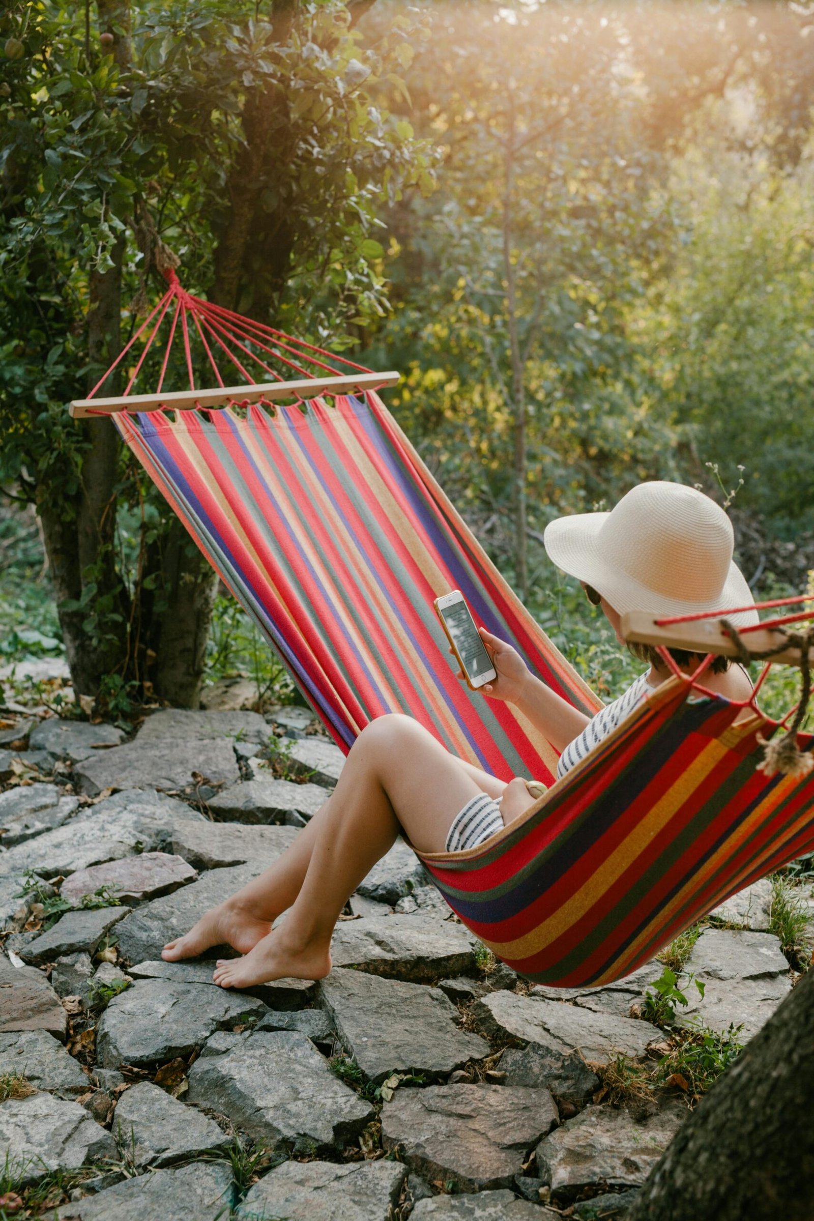 A woman enjoys a tranquil moment in a hammock using her smartphone in a sunny garden.