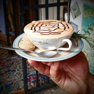 Mano sosteniendo una taza de café con arte latte en la espuma, servida en una taza antigua con un plato, una galleta y una rama de romero.