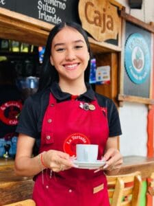 Mujer sonriente con uniforme de café ofreciendo una taza de café en La Tertulia Café, Quindío