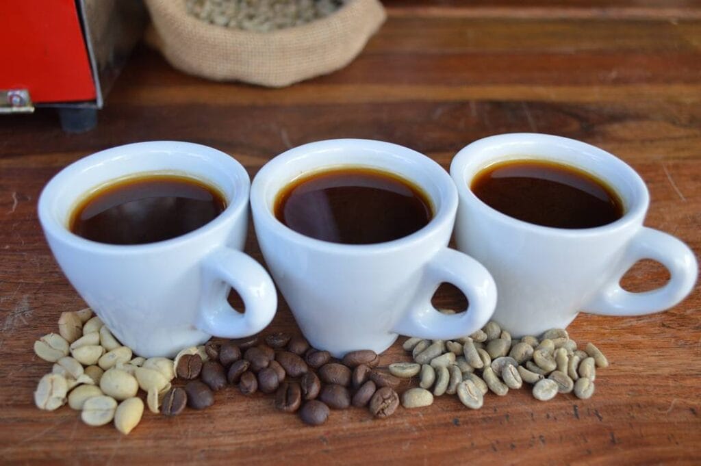 Tres tazas de café y granos de café de diferentes variedades, verdes y tostados, sobre una mesa de madera