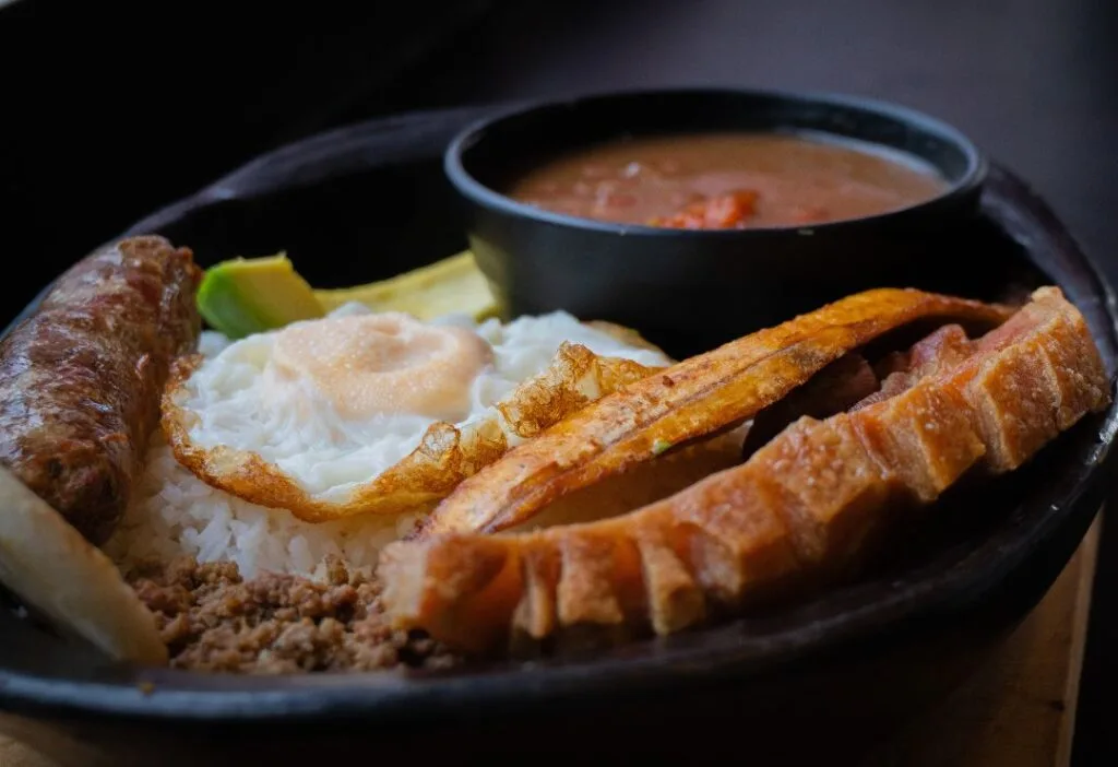 Primer plano de una auténtica Bandeja Paisa colombiana en un plato de barro negro, con arroz, frijoles, carne molida, chicharrón crocante, huevo frito y chorizo.