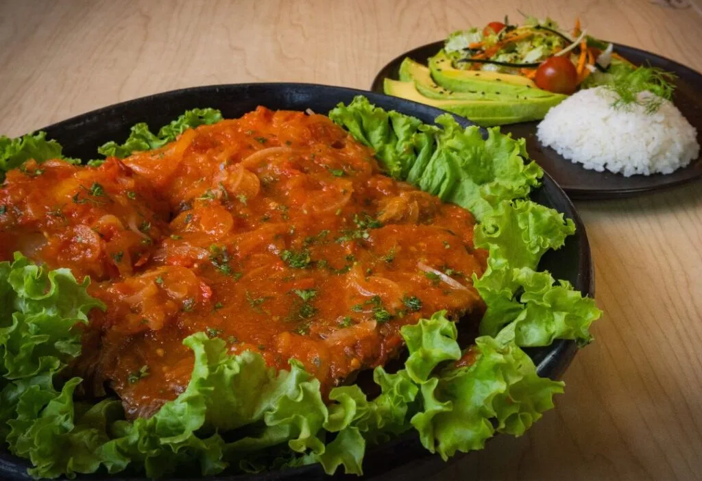 Dos platos individuales sobre una mesa de madera, mostrando un filete cubierto con salsa criolla (hogao) sobre lechuga y un plato de acompañamiento con arroz y ensalada de aguacate