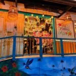 Vista de la entrada del Restaurante La Martina, mostrando una colorida pared exterior con un mural paisajístico, una baranda azul y la decoración floral y luminosa del interior visible a través del acceso