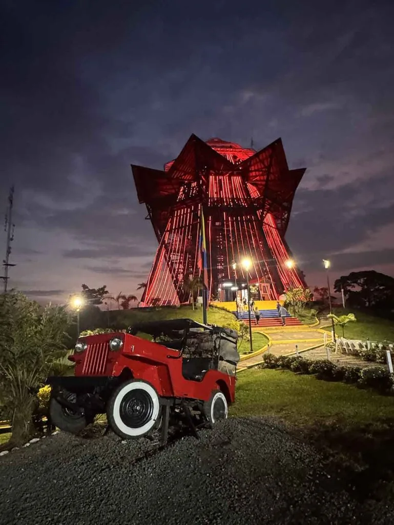 Fotografía nocturna del Mirador Colina Iluminada en Filandia, Quindío, brillando con luces rojas intensas. En primer plano, sobre una base rústica de grava, un Jeep Willys clásico de color rojo. Un sendero iluminado con faroles conduce a la torre bajo un cielo nublado al anochecer.