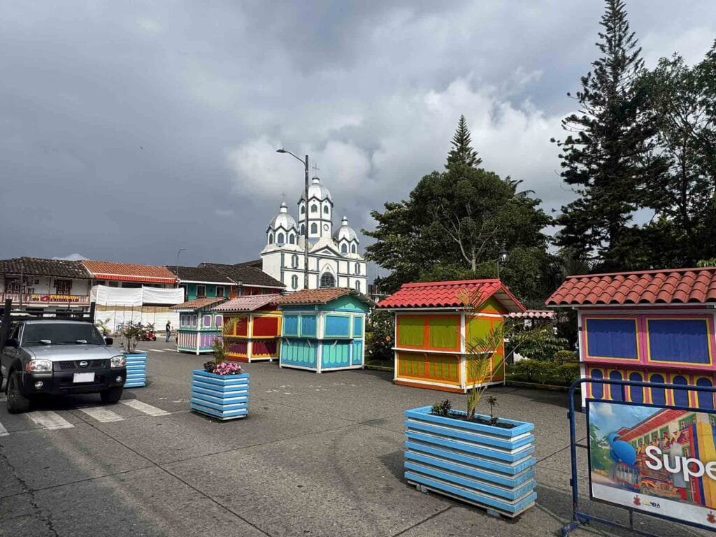 Plaza principal de Filandia, Quindío, con puestos de artesanías coloridos, maceteros azules y la Parroquia María Inmaculada al fondo bajo un cielo nublado.